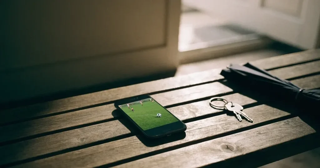 Ein Schlüsselbund und ein Smartphone mit dunklem Display liegen auf einer Holzbank in einem Hauseingang, daneben ein Regenschirm.