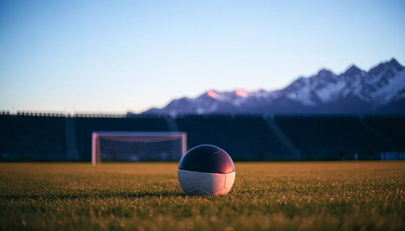Ein Fussball liegt auf dem Rasen eines leeren Stadions am frühen Abend, dahinter erheben sich schneebedeckte Berge unter klarem Himmel.