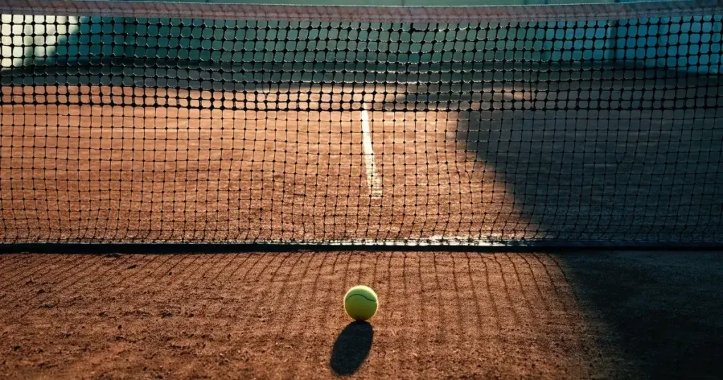 Ein Tennisplatz im ersten Morgenlicht, das Netz hängt leicht durch und auf der Grundlinie liegt ein einzelner Ball.