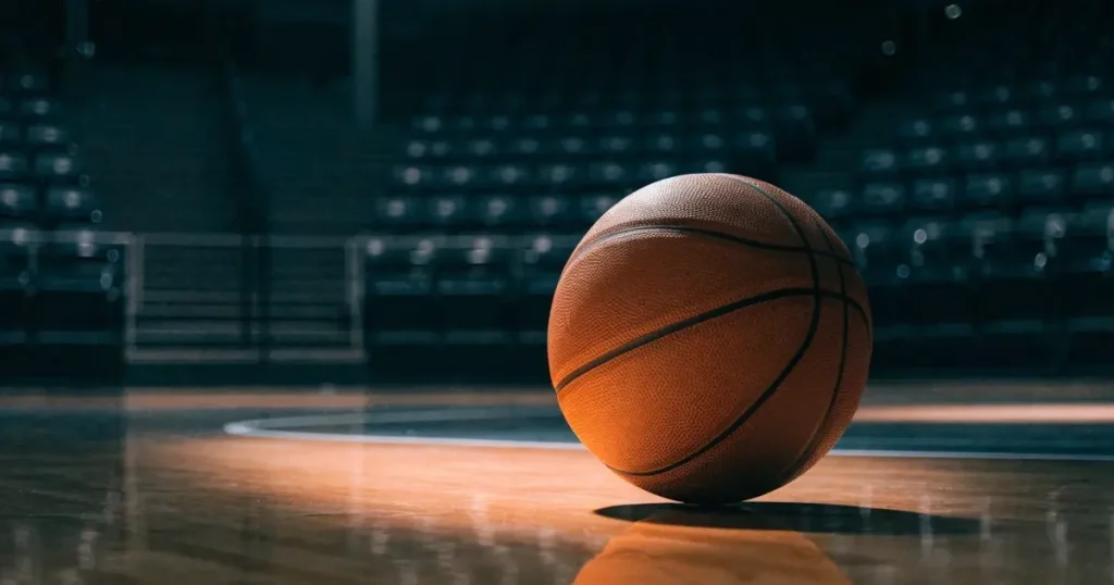 Ein Basketball liegt auf dem hellen Parkett einer leeren Halle, im Hintergrund steigen die Tribünenreihen im Halbdunkel an.