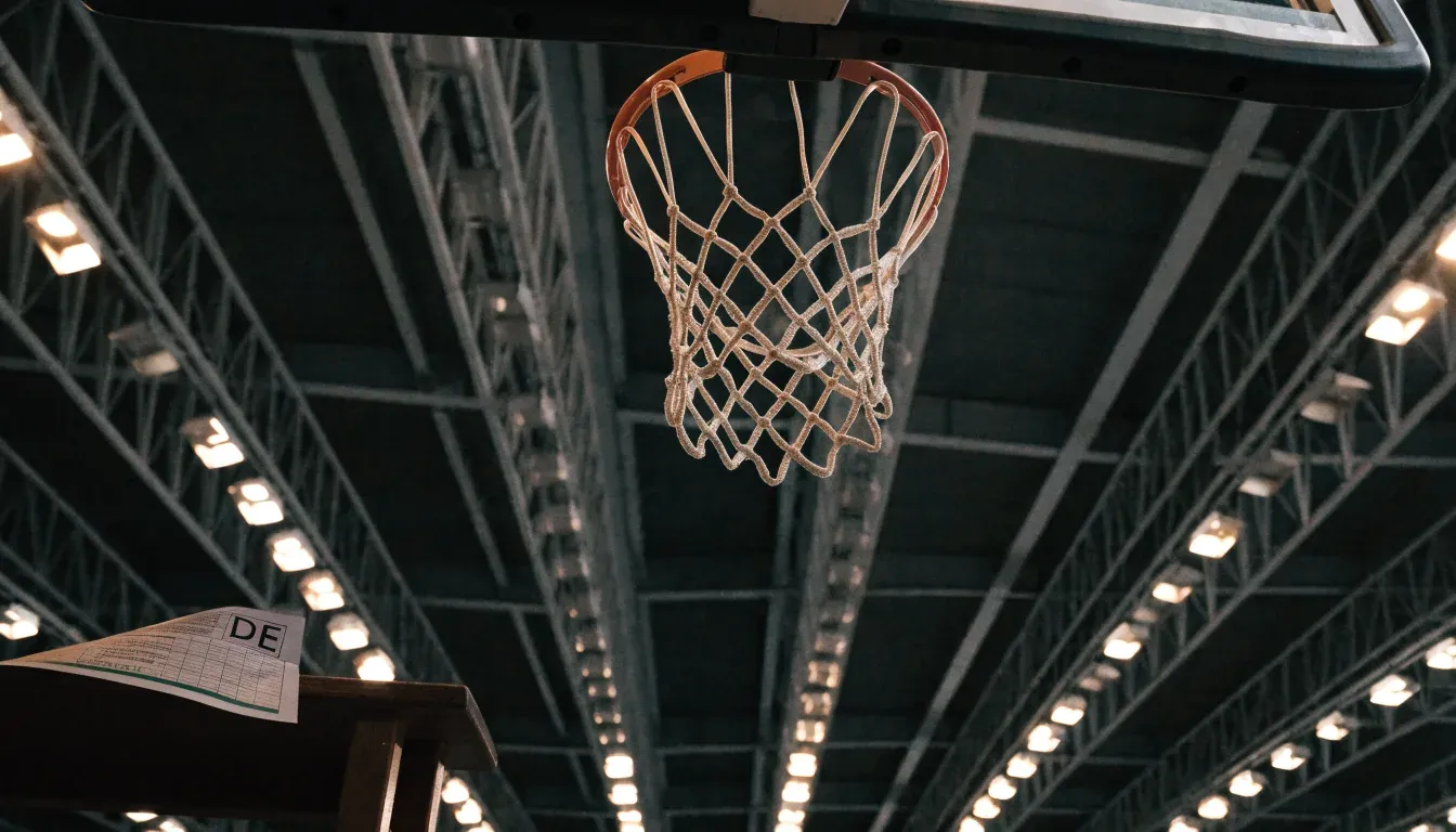 Ein Basketballkorb von unten gesehen, darüber eine Hallendecke mit Scheinwerfern und Stahlträgern.