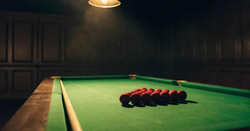 Ein Snookertisch mit grünem Tuch und einem Dreieck aus roten Kugeln unter einer tief hängenden Lampe in einer leeren Halle.